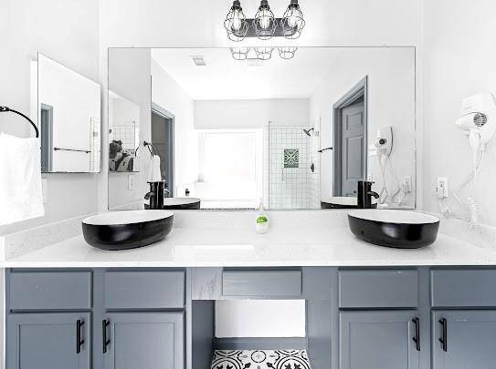 This bathroom features a modern design with dual sinks set in a white countertop, stylish gray cabinets, and a large mirror. The space is bright with sleek black fixtures, a geometric floor pattern, and glass-enclosed shower.