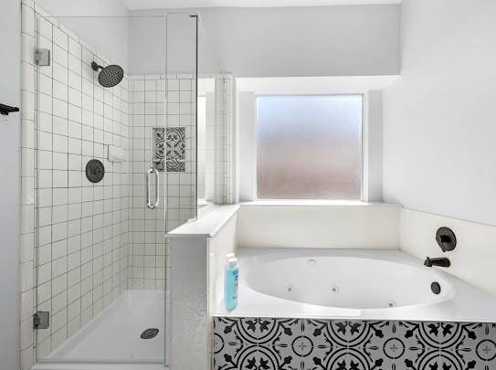 This bathroom features a modern glass-enclosed shower with white tile and decorative accents, alongside a luxurious whirlpool tub. It has a frosted window for natural light and privacy, with stylish black fixtures and patterned tile detail on the tub surround.