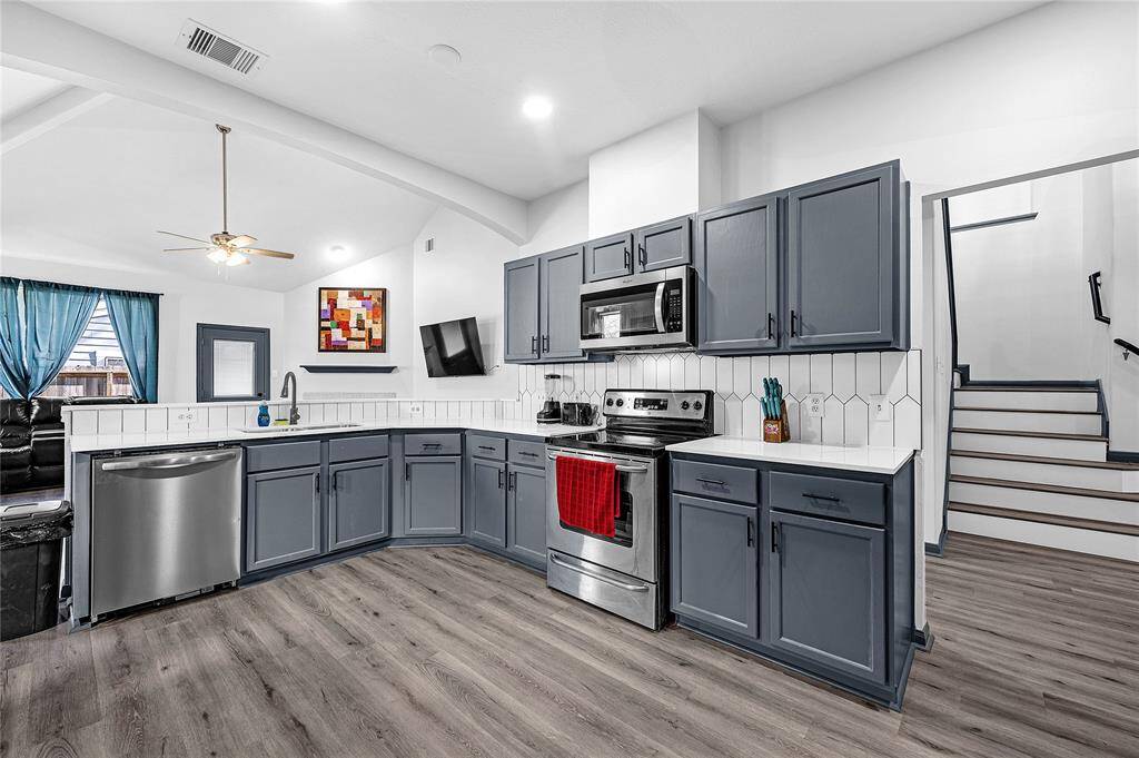 This kitchen features modern gray cabinets, stainless steel appliances, and a stylish hexagonal tile backsplash. It opens to a living area with a ceiling fan and natural light from large windows, creating a bright and inviting space.