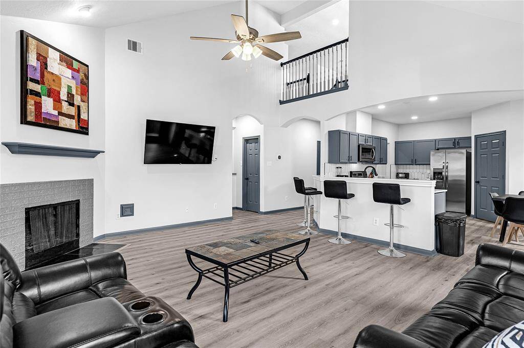 This spacious living area features an open floor plan with modern finishes, including a sleek kitchen with blue cabinetry, a cozy fireplace, and high ceilings with a ceiling fan. The room is well-lit, has a mounted TV, and offers a stylish, contemporary feel with laminate flooring and a bar seating area.