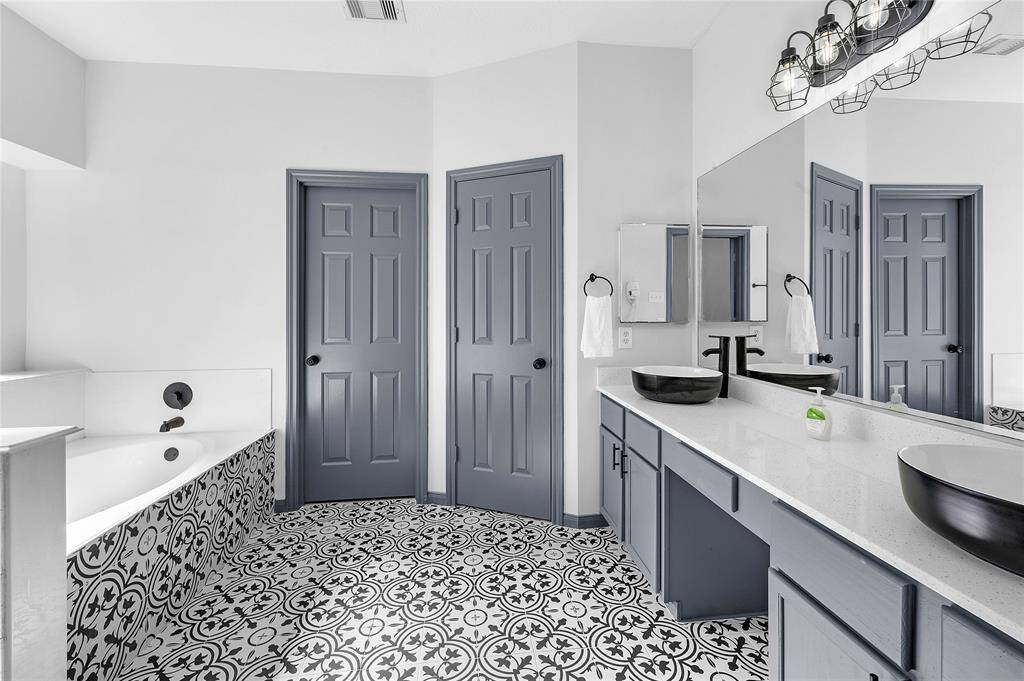This bathroom features a modern design with patterned black-and-white tile flooring, dual sinks with sleek black vessel basins, and ample counter space. The bathtub is accented with matching tile, and the room is finished with soft gray cabinetry and doors for a contemporary look.