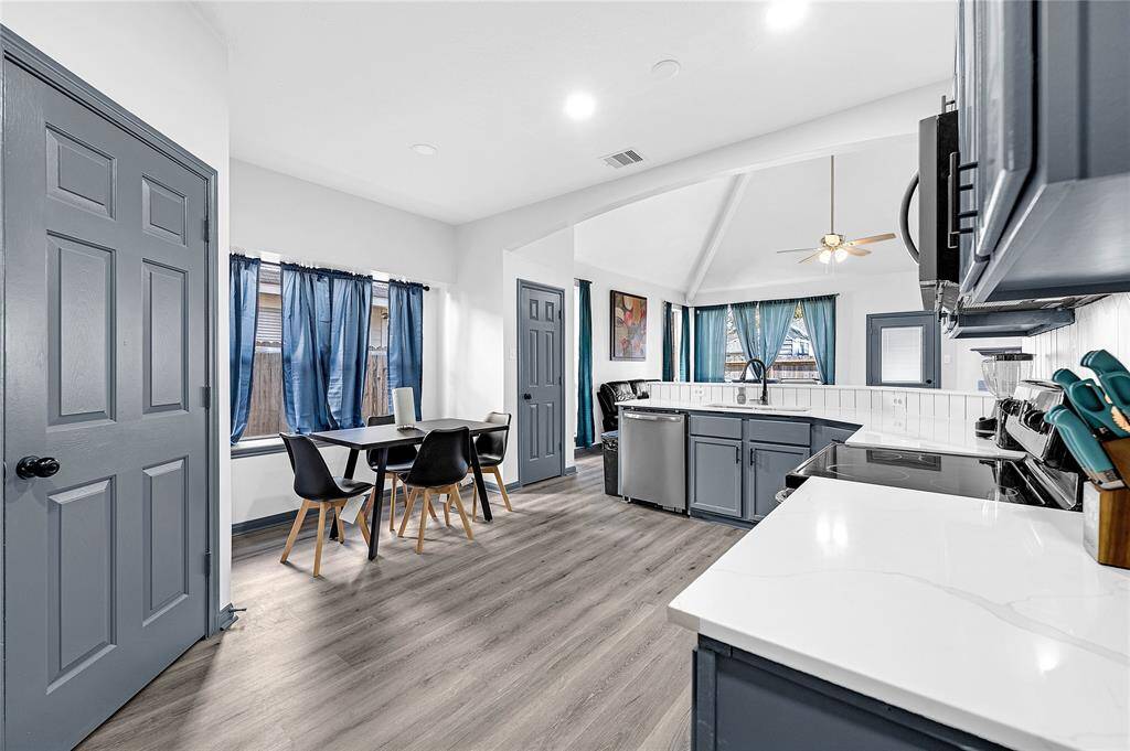 This photo showcases a modern kitchen and dining area with sleek gray cabinets, white countertops, and stainless steel appliances. The space is brightened by natural light through large windows with blue curtains. The open layout features a ceiling fan and light wood flooring, creating a welcoming and functional environment.