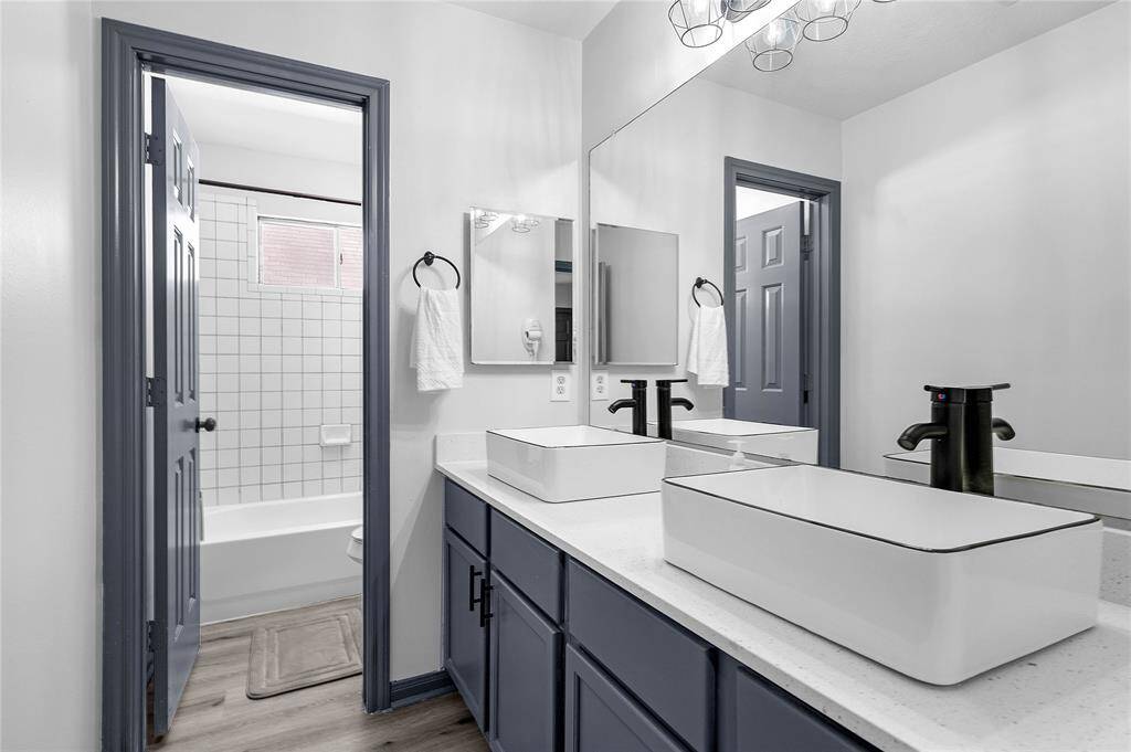 Modern bathroom featuring a double vanity with sleek vessel sinks and black fixtures. It has a large mirror, stylish lighting, and an adjacent bathtub with a tiled wall. The cabinetry and trim are in a dark, contemporary color, complementing the light walls and flooring.