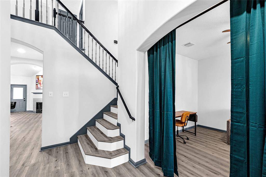 This photo showcases a bright, modern entryway featuring a staircase with dark railings, light wood floors, and an adjacent room separated by teal curtains. The space has a clean, contemporary look with neutral walls and ample natural light.