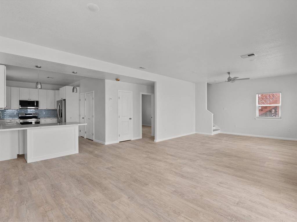 This photo showcases a spacious, open-concept living area with light wood flooring, a modern kitchen featuring white cabinetry and stainless steel appliances, and a small staircase leading upstairs. Natural light fills the space through a window, creating a bright and inviting atmosphere.