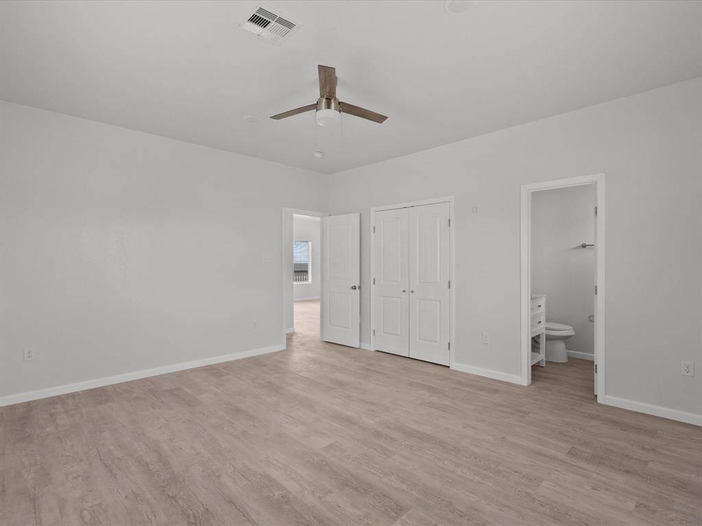 This bright bedroom features light wood flooring, neutral walls, a ceiling fan, and ample closet space. It includes an ensuite bathroom and connects to another room, providing convenience and functionality.