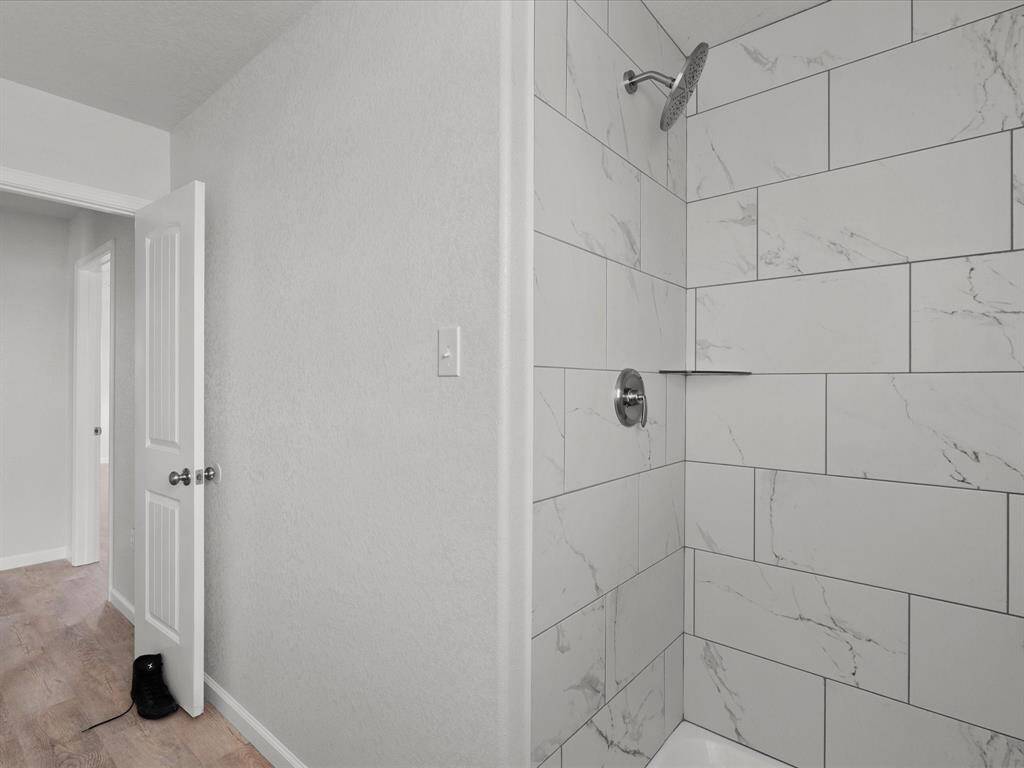 Modern bathroom with a sleek, tiled shower featuring marble-like tiles and chrome fixtures. The space is bright with light-colored walls and wood flooring visible in the adjacent hallway.