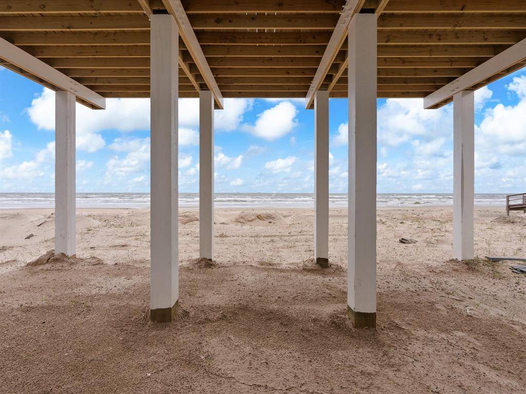 This photo shows the underside of a beachfront house with sturdy white pillars on sandy terrain, offering direct access to the beach and stunning ocean views under a blue sky with scattered clouds.