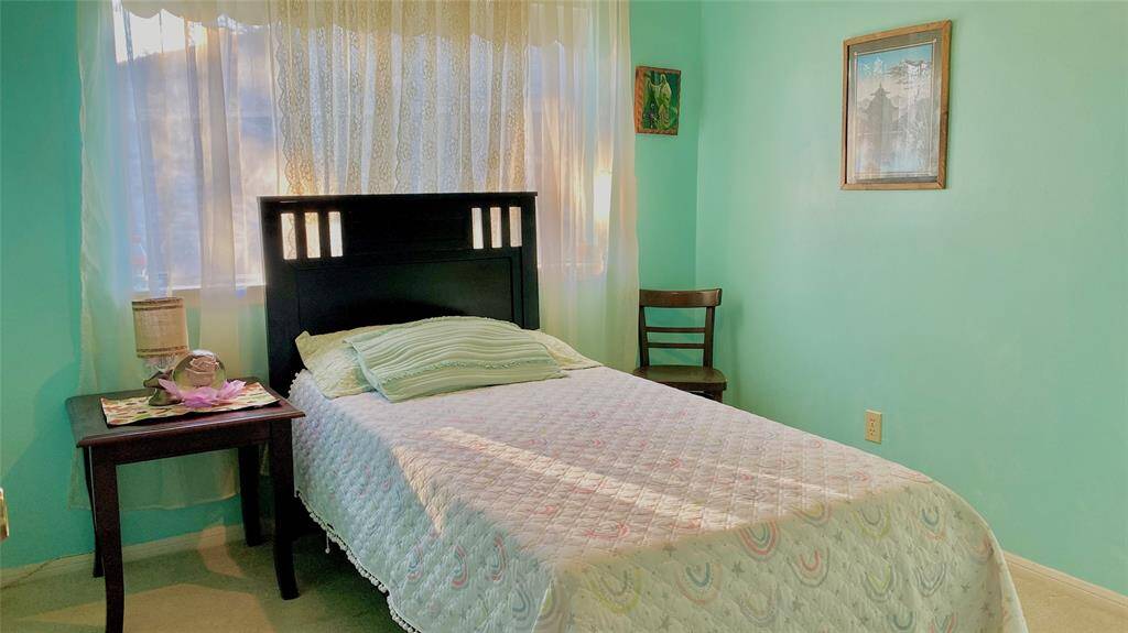 This cozy bedroom features a single bed with a dark wooden headboard, soft green walls, and a large window with sheer curtains allowing natural light. A small bedside table and chair add functionality to the space.
