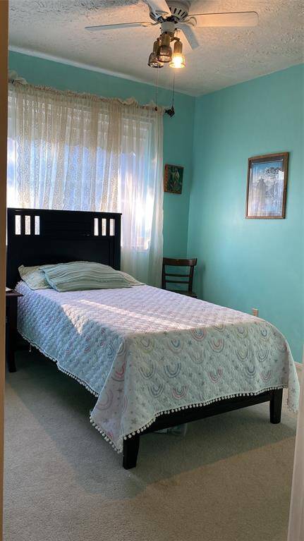 This cozy bedroom features a single bed with a decorative quilt, a ceiling fan, and soft teal walls. Natural light streams through sheer curtains, creating a bright and inviting atmosphere.