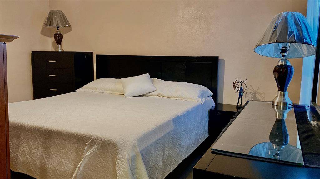 A cozy bedroom featuring a comfortable double bed with a white quilt, complemented by a sleek black headboard and matching bedside tables with lamps. The room has a modern, minimalistic feel with neutral walls and ample storage in a black dresser.