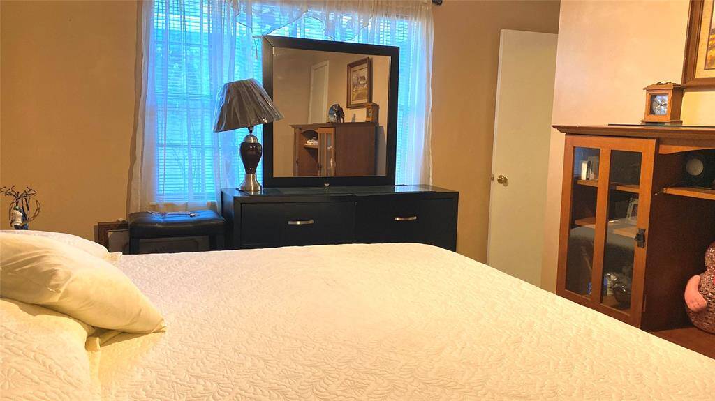 This cozy bedroom features a large window with sheer curtains, allowing natural light to brighten the space. It includes a comfortable bed, a sleek black dresser with a mirror, and a wooden cabinet for storage. The room has a warm, inviting atmosphere.