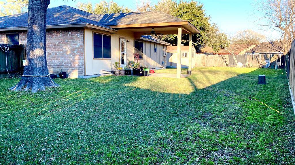 This backyard features a spacious lawn with a covered patio area attached to a brick and siding house. The yard is fenced, offering privacy, and there's a large tree providing shade. Ideal for outdoor activities and relaxation.