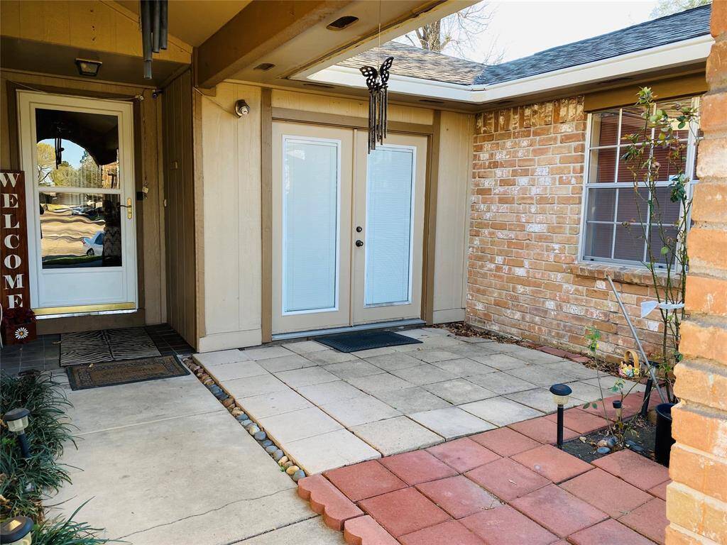 This welcoming entryway features a covered porch with a mix of brick and siding, double glass doors, and a small paved patio area. It has a charming atmosphere with a wind chime and a "Welcome" sign by the door.