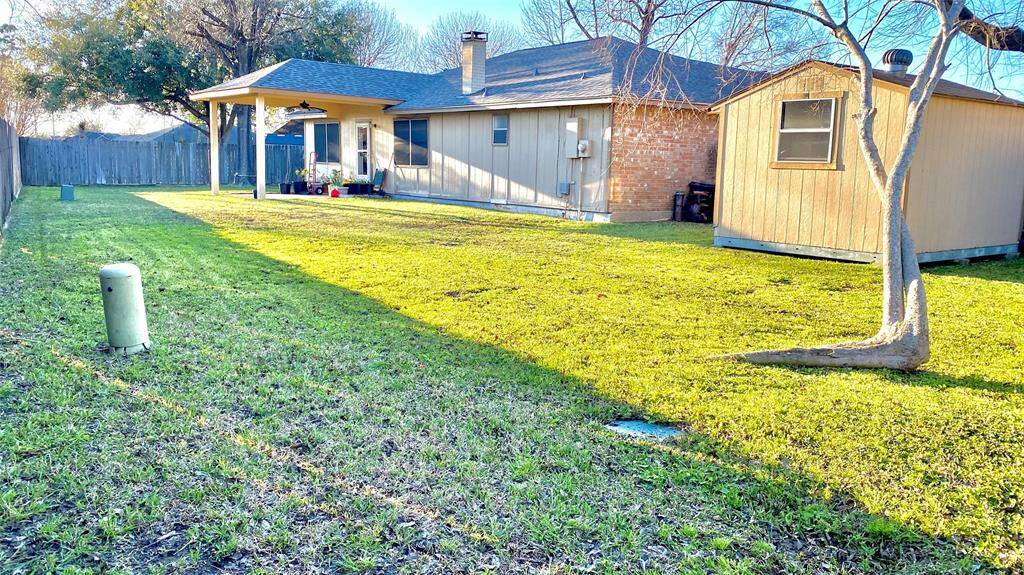 This photo shows a spacious backyard with a lush green lawn, a patio area, and a storage shed. The house has a brick exterior and a covered porch, offering plenty of outdoor living space.