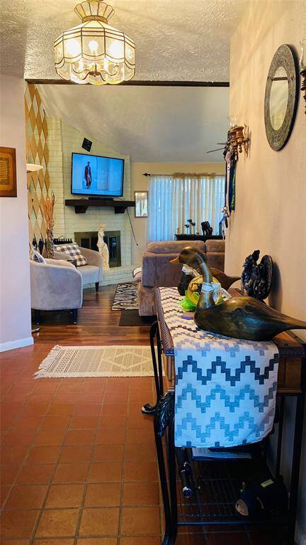 This cozy living room features warm wood floors and a brick fireplace with a mounted TV above. The space is well-lit with a large window and has inviting seating. Decorative elements include a stylish chandelier and unique wall art, creating a welcoming atmosphere.
