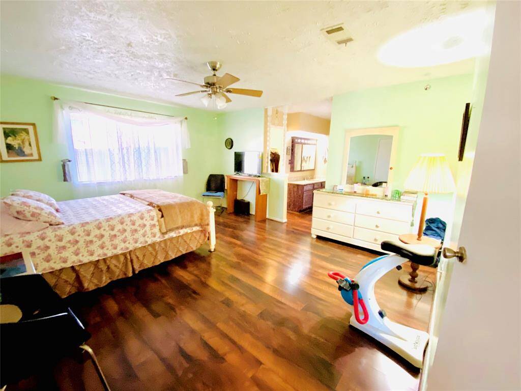 This spacious bedroom features hardwood floors, a ceiling fan, and ample natural light from a large window. It includes a cozy bed, a dresser with a mirror, and an exercise bike, creating a comfortable and functional living space.