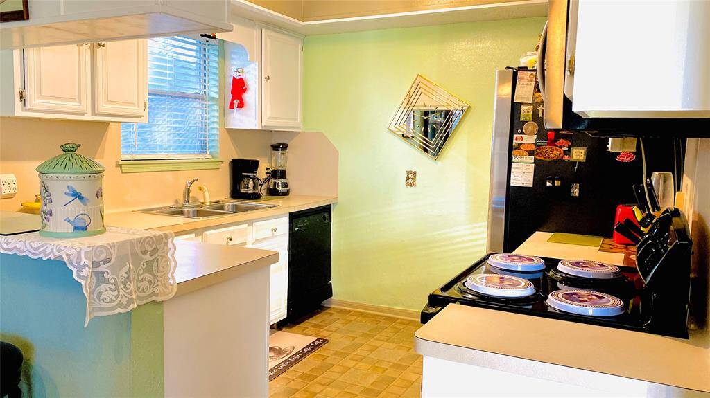 This kitchen features bright white cabinetry, a dual sink, and modern appliances, including a black refrigerator and stove. It has a cozy layout with a window providing natural light and a unique decorative mirror on a light green wall. The countertop space is accented with a lace runner and decorative jar.
