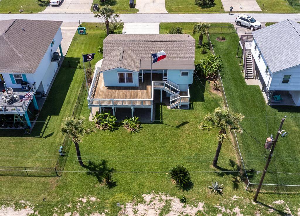 Charming coastal home with a spacious wooden deck, well-maintained lawn, and palm trees. It has a raised structure with stairs leading to the entrance, nestled between similar homes in a quiet neighborhood. Perfect for enjoying outdoor living and entertaining.