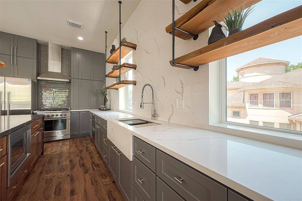 This is a modern kitchen with sleek gray cabinetry, stainless steel appliances, white countertops, and a stylish tiled backsplash. It includes an open shelving unit and has rich, dark wood flooring. Large windows allow for natural light.