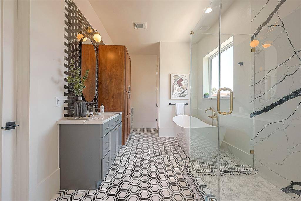 This is a modern bathroom featuring a geometric patterned floor, a sleek standalone bathtub, a glass-enclosed shower with marble walls, a vanity with gray cabinetry and a wooden storage cabinet, accented by gold-toned fixtures and a round mirror.