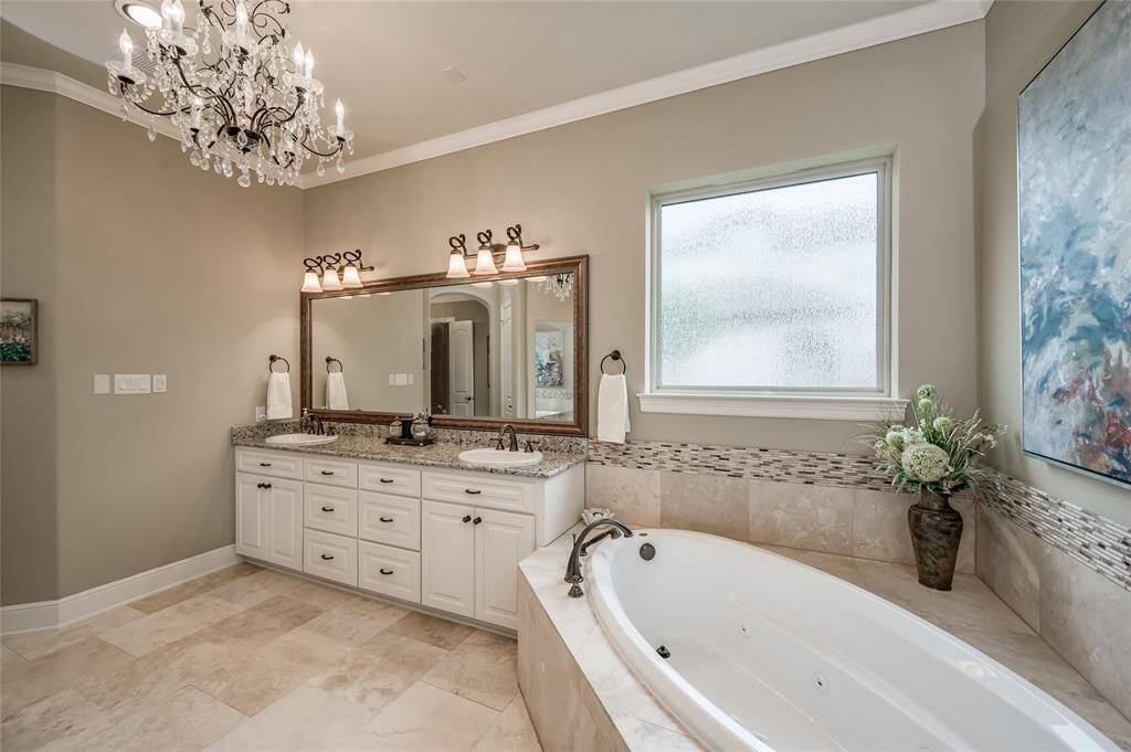 Ample natural light fills the space, highlighting the character of honed travertine stone. A large framed mirror sits atop granite counters with dual sinks.