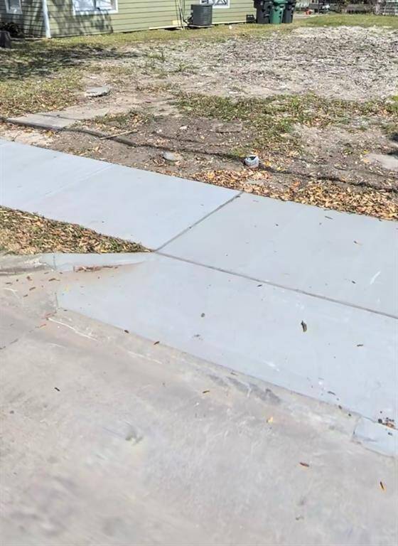 This photo shows a concrete driveway leading up to a modest home with a yard. The surrounding area appears to be in a suburban setting with some greenery and a residential structure in the background.
