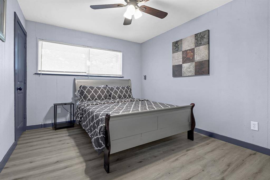 Discover a serene minimalist bedroom featuring soothing blue walls, elegant wood flooring, and a chic patterned bedspread. Enjoy the modern ceiling fan, sleek small table, and striking abstract wall art.