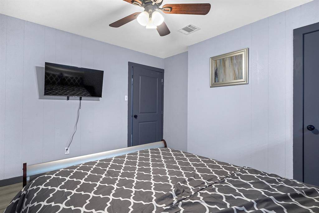 Inviting bedroom featuring soothing light blue walls, a wall-mounted TV, a stylish patterned bedspread, a ceiling fan, and elegant dark doors.