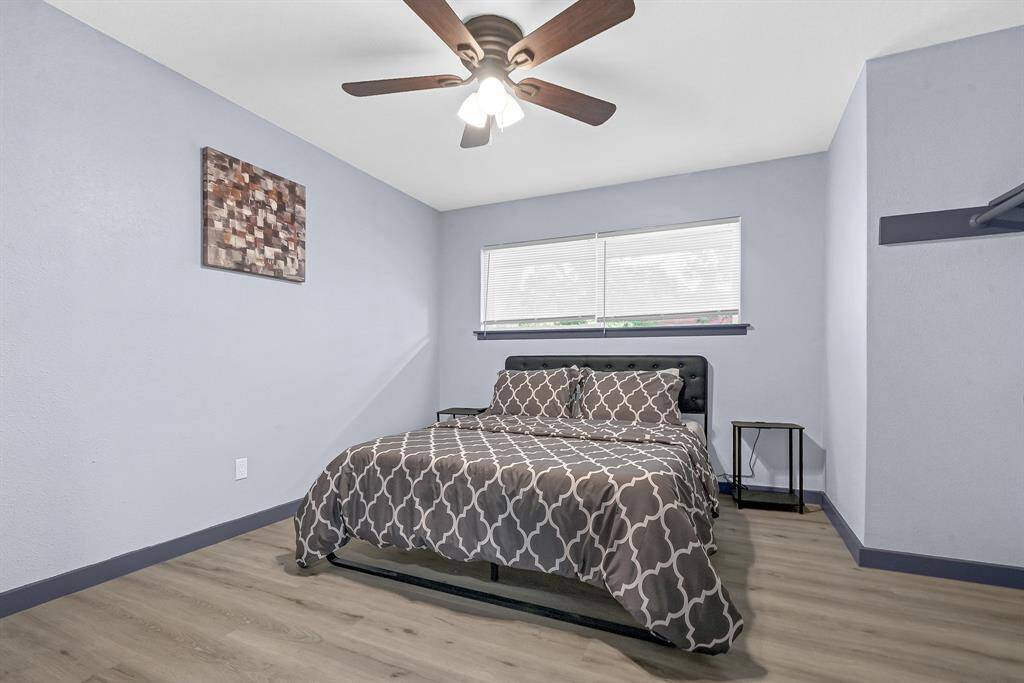 Discover a cozy bedroom featuring elegant wooden flooring, soothing blue walls, and a stylish patterned bedspread. Enjoy the comfort of a ceiling fan and natural light through a window with blinds.