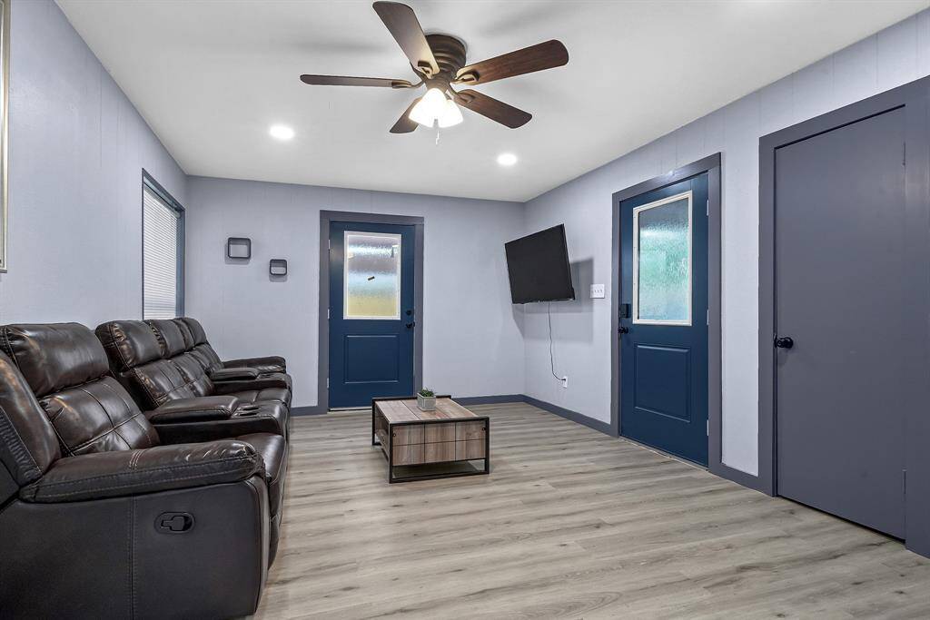 Experience modern comfort in this living room, featuring a sleek leather couch, elegant wooden floors, vibrant blue doors, a ceiling fan, and a wall-mounted TV.