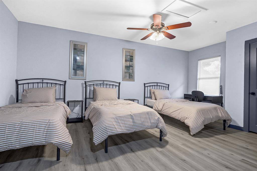 Discover a stylish, serene bedroom featuring three striped beds, elegant wooden flooring, and soothing blue walls, complemented by a ceiling fan for comfort.