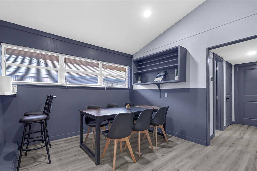 Discover a modern dining room featuring elegant dark blue walls, a stylish wooden table, sleek chairs, and bar stools. Enjoy ample storage with built-in shelves and abundant natural light through large windows with blinds.
