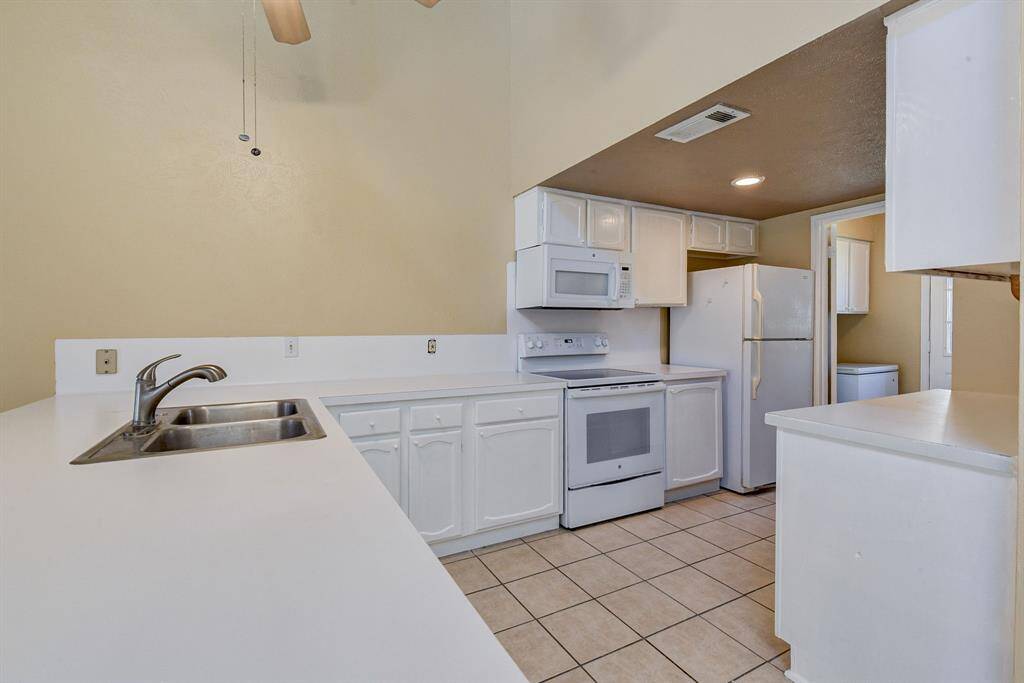 This kitchen features clean white cabinetry and appliances, a spacious countertop with a double sink, and neutral tile flooring. It offers a bright and functional space.