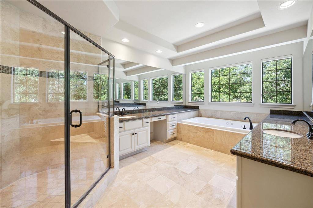 The primary ensuite bathroom boasts stone-tiled flooring, dual sinks atop granite countertops, a spacious jetted tub, vanity space, large shower and windows that bathe the room in natural light.