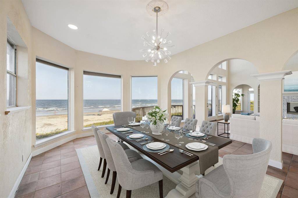 A coastal dining room with a beach view, elegant table setting, arched doorways, and a modern chandelier. This open area dining room is virtually staged and is 20 feet by 12 feet long with the breathtaking view of the water.