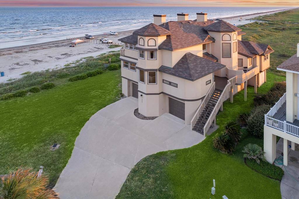 The photo showcases a luxurious multi-story beachfront property with a sweeping lawn, direct beach access, and multiple balconies offering panoramic ocean views.