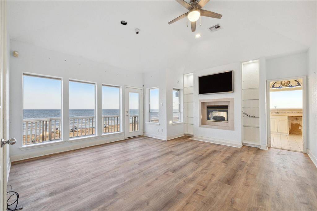 Bright beachfront room with large windows, a fireplace, a mounted TV, and wooden flooring leading to an outdoor deck with ocean views. This is one of the 2 primary bedrooms.