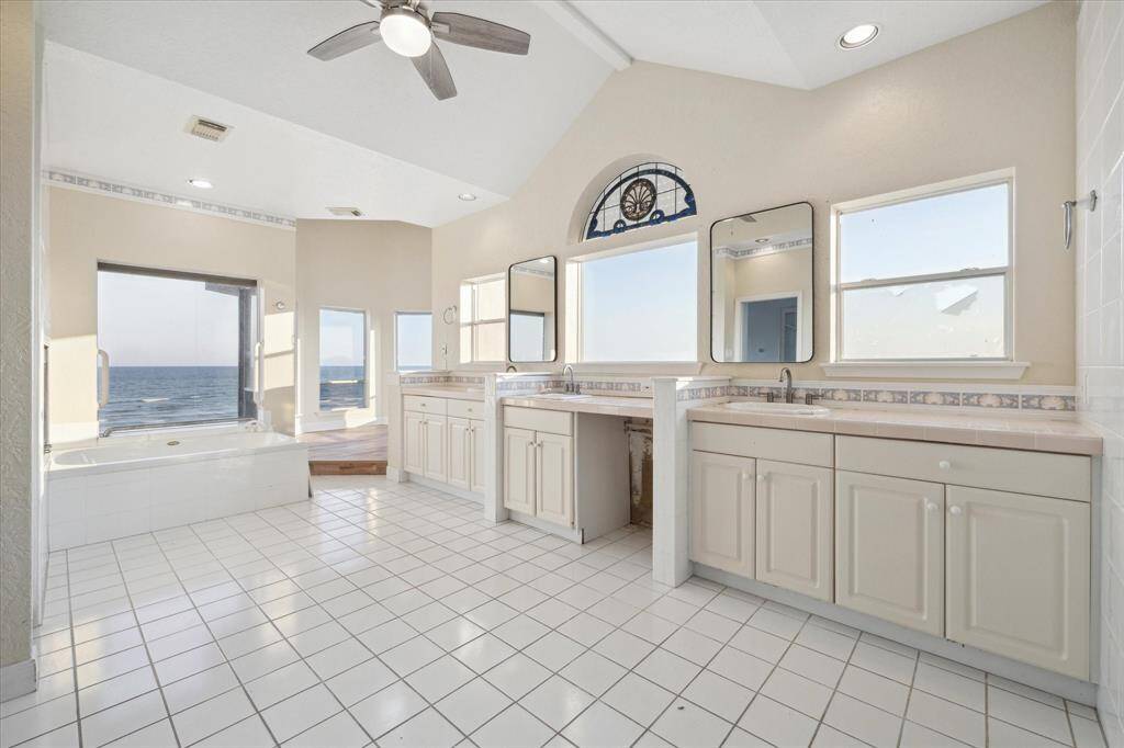 Bright, spacious bathroom with ocean view, jacuzzi tub, dual sinks, oversized mirrors, tiled floor, and nautical-themed clock above the window. There is a place to have your exercise equipment or a sitting area to see ocean views and exercise.