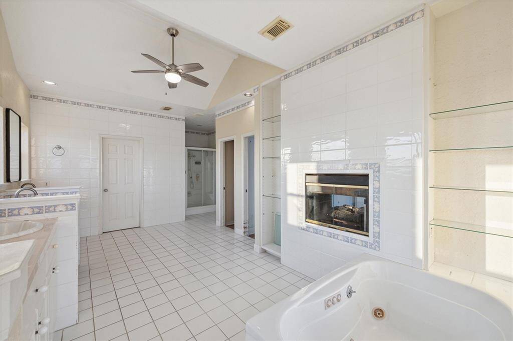 This spacious, bright bathroom has an open floor, a corner tub, a white vanity, glass shelves, a ceiling fan, and a fireplace. There is also a bidet and two toilets for your convenience.