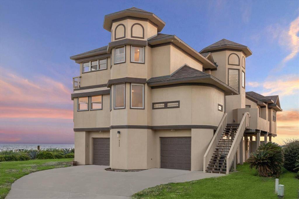 This is a multi-story, modern beachfront home with a unique architectural design, featuring multiple balconies, large windows, and a two-car garage. The property enjoys an elevated position with a lush lawn and boasts impressive sunset views over the ocean horizon.