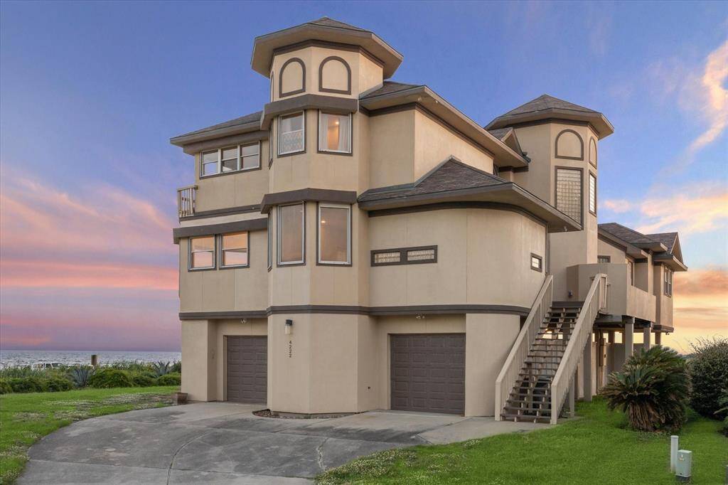 Multi-story beige house with multiple garages and balconies, set against a sunset sky, adjacent to a coastal area with greenery.