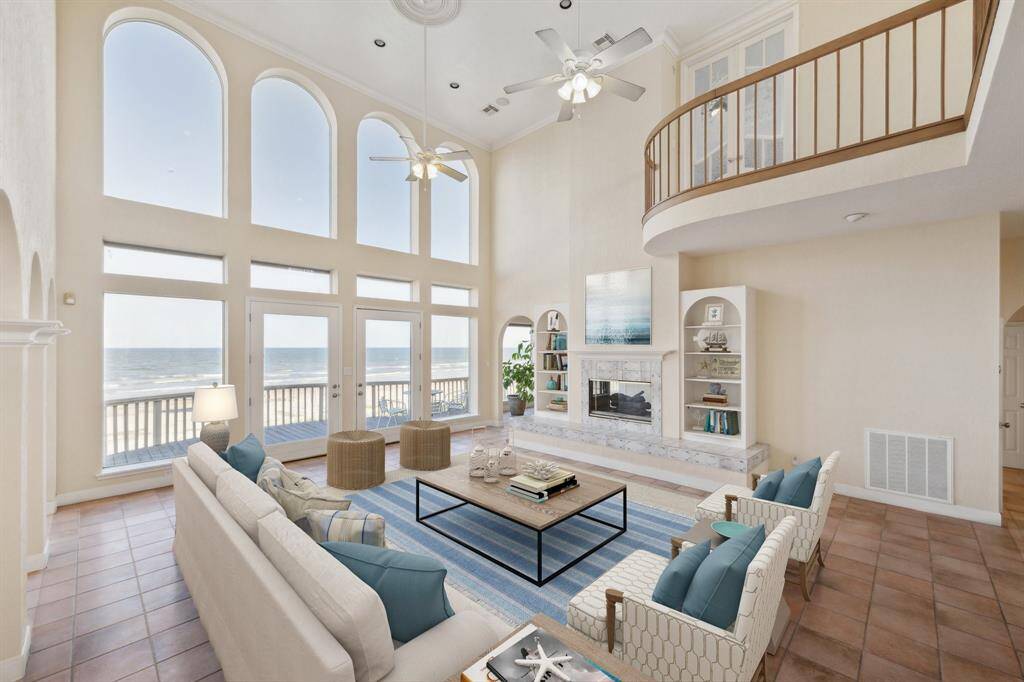 A luxurious beachfront living room with high ceilings, tall windows, elegant furnishings, and a balcony overlooking the sea. This is a virtually stage room.