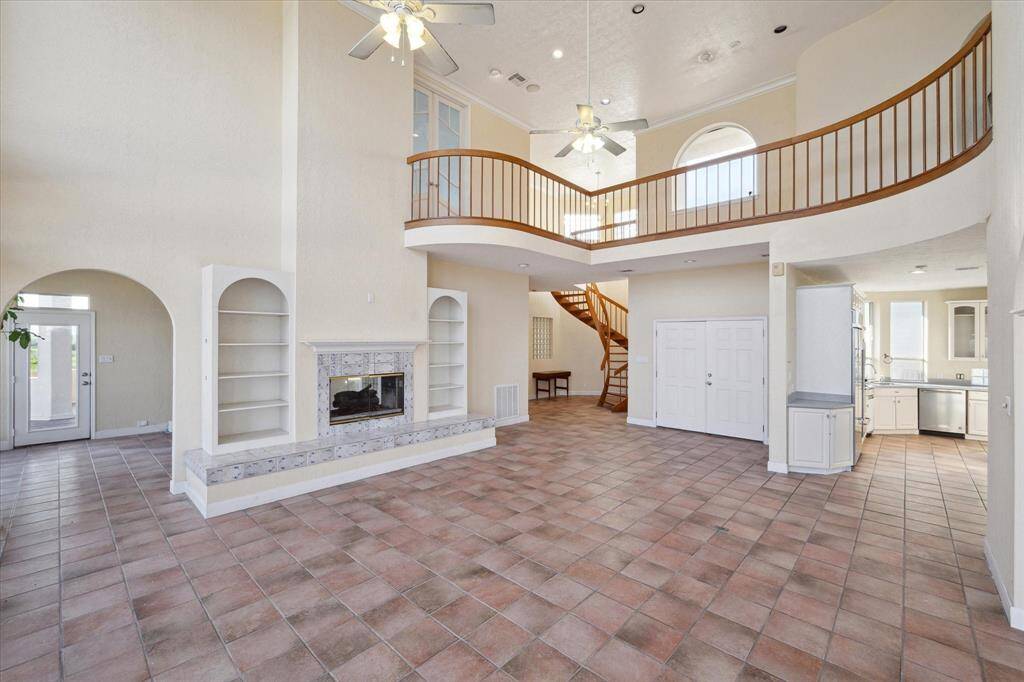 A spacious living room with high ceilings, a fireplace, tiled floor, and an open staircase leading to a second-floor balcony with elevator.