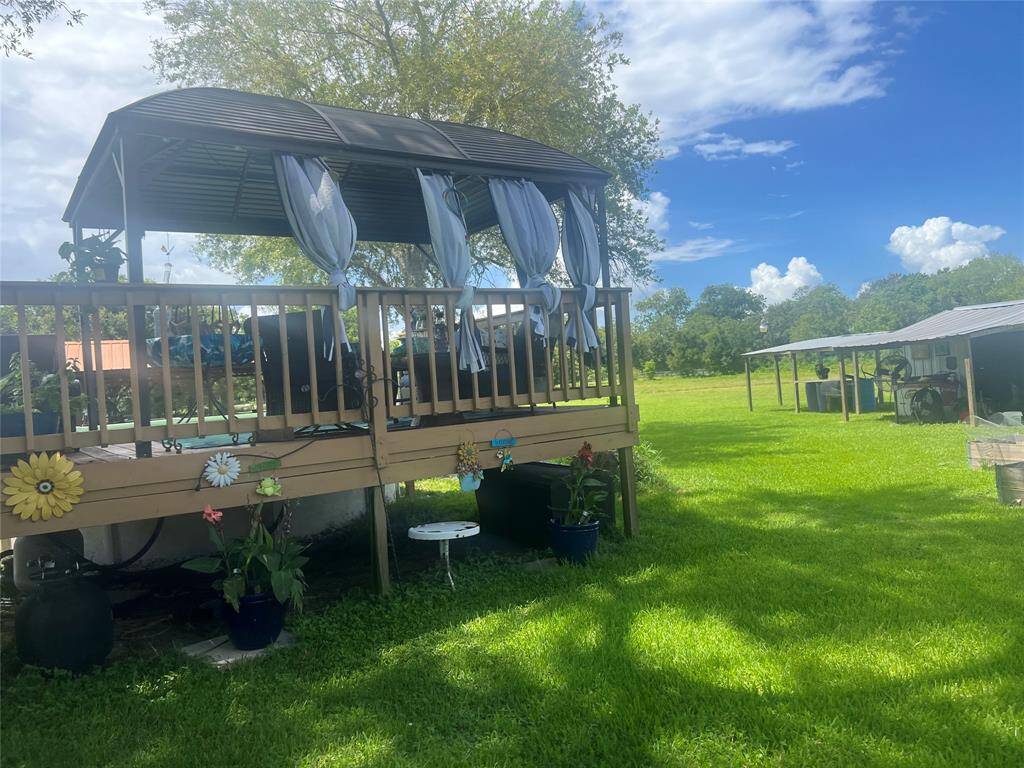Another charming outdoor space featuring a cozy, covered deck adorned with sheer curtains and decorative elements, overlooking a spacious, well-maintained lawn with ample greenery and a clear, sunny sky. There's a carport visible in the background, suggesting additional utility and storage space.
