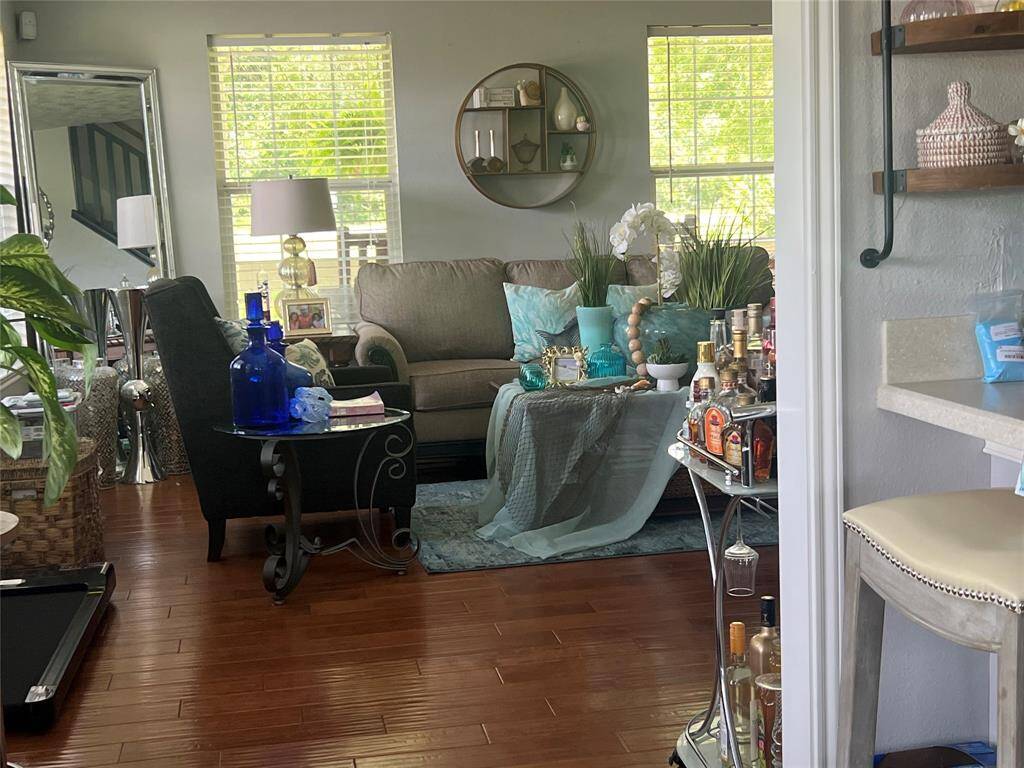 This is a cozy, well-lit living space with hardwood floors and large windows with greenery views. The room features tasteful decor, comfortable seating, and a bar cart, creating a welcoming atmosphere for relaxation and entertainment.