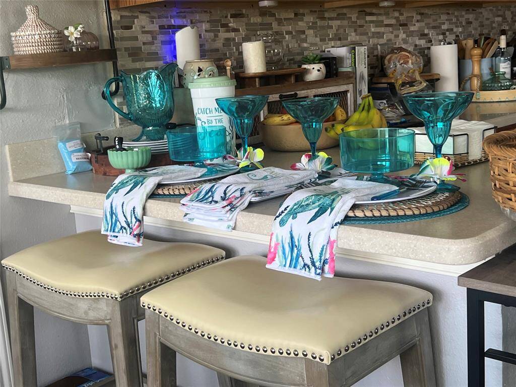This is a kitchen counter space with a breakfast bar setup, featuring two upholstered bar stools. The counter is styled with various decorative items in a coastal theme, including turquoise glassware and accent pieces. It has a mosaic tile backsplash that complements the overall aesthetic. Please note that the space is currently in a lived-in state with some personal items and kitchenware on display.