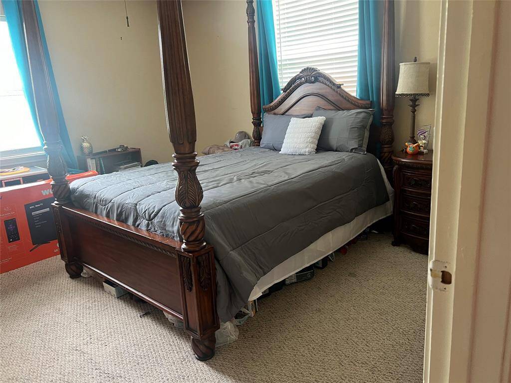 This is a well-sized bedroom featuring a large four-poster bed with coordinating bedside tables and a classic lamp, accentuated by contrasting teal window treatments. The room has neutral walls and carpeting, providing a warm and inviting space.