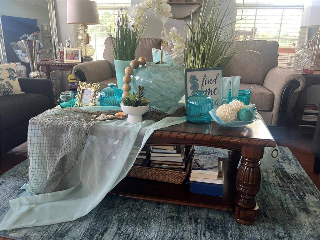 The living room has a nautical theme, featuring a wooden coffee table adorned with decorative items, surrounded by comfortable seating and natural light filtering through the blinds.