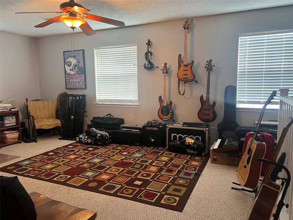 This is a spacious games room with neutral walls and carpet flooring, currently set up as a music studio with multiple guitars and equipment. The room includes a ceiling fan and natural light from two windows with blinds.